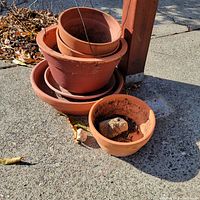 Photo showing a stack of seven terra cotta pots and one terra cotta saucer outdoors on concrete surface with autumn leaves nearby. Pots vary in size with soil and weathering visible.