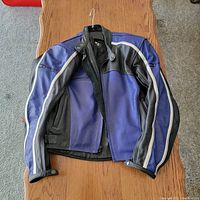 Front view of the leather motorcycle jacket showing blue and black panels with white stripes, laid flat on a wooden table with zipper open.