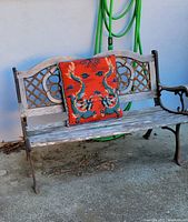 Outdoor wood bench with distressed seat and fleur de lys iron accents on armrests and backrest, shown with a red decorative cushion placed on seat.