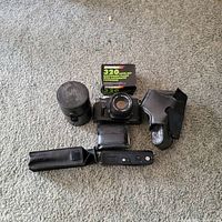 Top-down photo showing Ricoh camera with lens attached, lens case, Nikkor flash box, leather case, and accessory pouch arranged around it on carpeted floor.