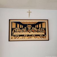 Full view of the framed tapestry depicting the Last Supper hanging on a white wall with a crucifix above it.