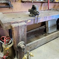 Close-up of thick wooden workbench leg with mounted metal vise handle and wood surface showing wear and distressing.