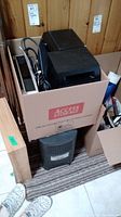 Box with multiple black audio components, including a media center and speaker system parts, cables visible.