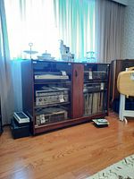 Wooden stereo cabinet with two glass doors letting view into compartments containing audio equipment and vinyl records, surfaces hold decorative items.