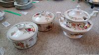 Three-piece set of ceramic serveware including soup tureen and two covered casseroles with floral and fruit decoration.