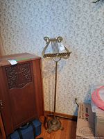 Full view of the brass music stand showing the ornate lyre-shaped holder, adjustable pole, and decorative base positioned in a corner by furniture and boxes.