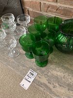 Photo showing green glass pitcher with six matching green glasses and six clear glasses with patterns on a stone surface near a brick wall.