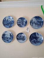 Six blue and white decorative wall plates hung on a wall. Each plate depicts detailed Dutch countryside and village scenes featuring horses, carts, windmills, and figures.