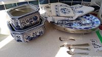 Photo showing 3 blue and white porcelain plant pots with floral patterns stacked, porcelain serving platter with metal base, folded white napkins with blue embroidery, and silver forks and spoons beside them on counter