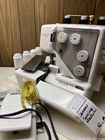 Front and side view of Janome MyLock sewing machine with foot pedal, measuring tape, and some white thread spools and accessories on the table.