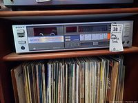 Full front view of Sony cassette deck model TC-FX66 with visible cassette compartment, control buttons, and analog meters. Shelf and vinyl records visible below.