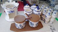 Photo showing two Blue Danube condiment jars with wooden tops and spoons set on a wooden tray, alongside nine matching ceramic spice jars with lids and a ceramic warmer with an opening for candle placement.