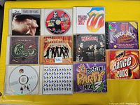 Photo of 11 music CDs laid out on a yellow fabric background showing covers featuring artists Tears for Fears, Judas Priest, Rolling Stones, Chicago, Spice Girls, Hedley, The Police and various music compilations.