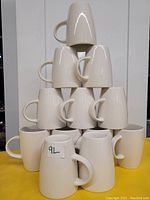 Photo showing 12 white ceramic mugs stacked in a pyramid formation against a plain background. The mugs have a smooth white finish and C-shaped handles. One mug features a small label marked '9L'.