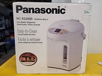 Photo of box showing front label and product image of Panasonic NC-EG3000 hot water dispenser, white color. Box mentions easy-to-clean charcoal coated non-stick interior and 3.0L capacity.