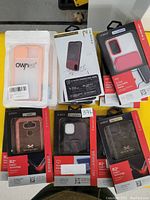 Photo showing multiple phone cases in packaging on a yellow surface, including Ownest and Xeotech branded cases, various colors and styles, demonstrating assortment and condition as new.
