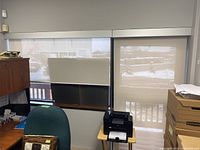 View of two rolling translucent blinds inside an office space, one covering a window approximately 68.5 x 52 inches and the other a door about 39 x 80 inches. Shows blinds partially rolled down with visible pull chains and mounted headrails.