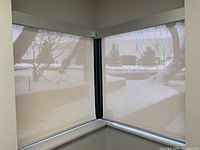 Two beige/off-white light filtering roller blinds installed at adjacent windows in a corner with snowy landscape visible outside. Both blinds have metal mounting rails and pull chains.