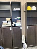 Tall faux wood bookshelf filled with books and papers, showing three shelves and a two-door cabinet below.