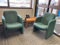 Pair of green upholstered armchairs and faux wood side table in a room near window with carpeted floor