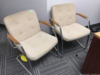 Pair of mid-century style chairs showing textured upholstery and tubular metal frames with wooden armrests, placed on office carpet.