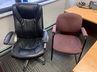 Two office chairs placed side by side on carpet. Left chair is black vinyl executive on casters with padded armrests. Right chair is maroon fabric upholstered with metal frame and no casters.
