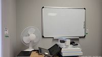 Photo shows 12-inch white desk fan placed on a small surface near a whiteboard mounted on a wall above it, also visible are some miscellaneous items near the fan.