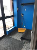 Entryway corner with four floor protector mats arranged on floor, yellow snow shovel leaning against wall, and black boot tray rolled up.