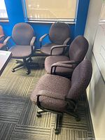 Photo showing four matching upholstered office chairs arranged near windows and walls, highlighting the fabric, armrests, and caster bases.