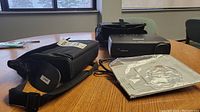 Projector and accessories including carrying case, cables, and manual on a wooden table.