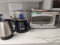 Photo of the three small appliances - Hamilton Beach electric kettle, PC coffee maker, and Oster toaster oven arranged on a kitchen counter with a white marble effect surface.