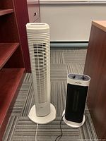 Front view of a white Sunbeam tower fan and a black/white Pelonis ceramic space heater on carpeted office floor.