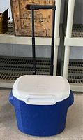 Full view of blue cooler showing white lid and black telescoping handle extended for rolling.
