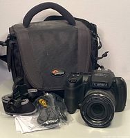 Fujifilm FinePix HS25EXR camera with lens, black and gray Lowepro camera bag behind, camera strap, cables in packaging, and user manuals stacked on table