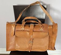 Front view of a brown handmade leather duffle bag resting on a chair, showing front flap pocket and buckle details.