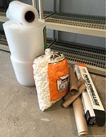 Photo showing two large rolls and one smaller roll of bubble wrap beside partial bag of packing peanuts and kraft paper rolls on a concrete floor in front of shelving