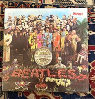 Front of the vinyl album cover showing The Beatles in colorful costumes surrounded by famous figures, with 'IMPORT' sticker on top right.
