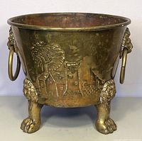 Front side view showing the embossed design of a house, tree, and wall with lion paw feet and a ring handle on the side.