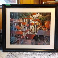 Framed print showing a vibrant small town scene with buildings and red roofs.
