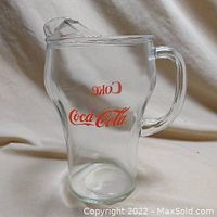 Front view of clear glass Coca-Cola pitcher showing red Coke and Coca-Cola logos and handle.