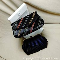 Two black clutch purses with one pleated fabric clutch framed in metal and one black beaded clutch with colorful diagonal stripes, posed on a beige fabric surface with a white Calvin Klein box behind them.