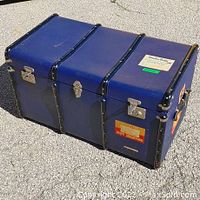 Photo of large blue steamer trunk with black reinforced edges, metal latches, and vintage travel stickers on top and side.