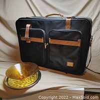 Black luggage bag with tan straps and handle, standing upright with front side facing camera, two large zippered pockets visible.