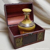 Glass decanter with gold decoration and stopper placed inside open wooden treasure chest with decorative contrasting panels.