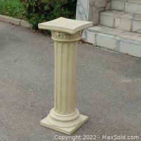 Front perspective view of the decorative classical column pillar placed outdoors on pavement near steps and greenery.