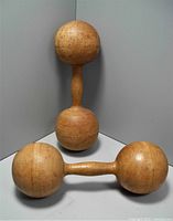 Two antique wooden dumbbells placed in an L-shaped corner. One dumbbell stands upright showing full height; the other lies horizontally showing spherical ends and handle.