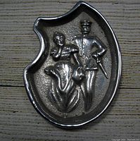 Front side of the aluminum pin tray showing embossed figures of a woman and a man in uniform.