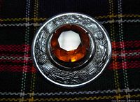 Close-up image showing the front of the round kilt pin with an amber-colored faceted glass stone held by metal prongs. The surrounding metal base features an engraved thistle design. Pin is positioned on tartan fabric.