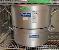 Two stacked round heavy gauge aluminum pots on a metal wire shelf. Both pots have side handles and Metalco branding labels on the front.