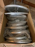 Top view of a box containing multiple stacked aluminum pot lids, showing their round metal shapes with handles.